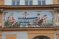 Ein alter Mann mit grauem Bart, Schurz und Krücke sitzt gemeinsam mit einer halbnackten mittelalten Frau und einem stehenden jungen, nackten Knaben um eine blaue Wanne, auf der ein Putto von weißen Tauben umflogen wird