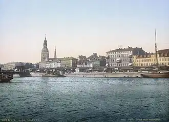 Alte Farbfotografie von einer Stadt mit der Aufschrift „Riga Le Quai“. Im Vordergrund ist ein breiter Fluss mit drei Schiffen. Am Ufer sind überdachte Stände und eine Brücke ist links teilweise zu sehen. Hinter der linken Häuserfassade sind zwei Kirchtürme.