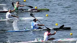 Vier Kajakfahrer bei einem olympischen Sprint-Rennen 2016 in Rio; die Sportler paddeln auf engem Raum nebeneinander, ihre Boote schneiden mit hoher Geschwindigkeit durch das Wasser. Die Kajaks tragen die Länderkennzeichnungen von Litauen, Ungarn, Italien und Frankreich.