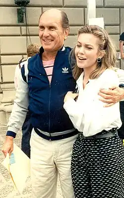 Diana Lane und Robert Duvall, die eng beieinander stehen und beide lächeln. Er auf der linken Seite trägt eine blaue Adidas-Jacke mit weißen und roten Streifen an den Ärmeln sowie ein rosa-weiß gestreiftes Shirt darunter. Sie hat eine helle Hose an. Diana Lane auf der rechten Seite trägt eine weiße Bluse und eine Hose mit einem schwarz-weißen Muster. Im Hintergrund erkennt man andere Menschen und ein Gebäude mit einer Fassade aus Stein. Das Bild vermittelt den Eindruck eines Alltagsmoments oder einer beiläufigen Begegnung.