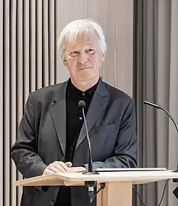 Älterer Mann mit grauen Haaren, schwarzem Hemd und grauem Anzug an Rednerpult