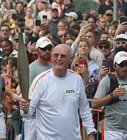Poirier trägt ein weißes Hemd und in der rechten Hand die olympische Fackel.