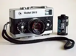 Frontale Farbfotografie einer analogen Kamera der Marke Rollei 35 S mit einer Schlaufe, die im Vordergrund liegt. Die Kamera hat neben dem Objektiv zwei Drehräder und auf der Kamera sind zwei Knöpfe und ein Spannhebel. Rechts steht eine Filmrolle.