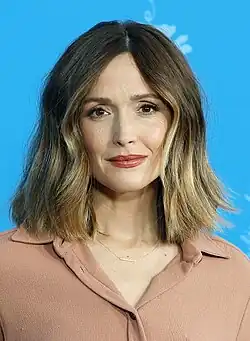 Rose Byrne auf der Berlinale 2025, mit schulterlangen, welligen Haaren und einem blassen Oberteil. Rose Byrne auf der Berlinale 2025 mit schulterlangem, welligem Haar, das dunkelbraun ist und helle Highlights aufweist. Sie lächelt in die Kamera und trägt eine hellbraune Bluse mit einem Kragen und eine zarte Halskette mit einem kleinen, horizontalen Anhänger. Der Hintergrund ist blau und mit weißen dekorativen Elementen gestaltet.