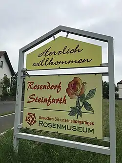 Ortseingangsschild vom Rosendorf Steinfurth heißt seine Gäste willkommen und ruft zum Besuch des einzigartigen Rosenmuseums auf.