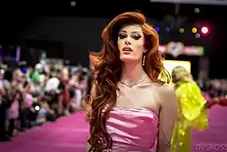 Porträtfoto von Goode, die auf einem roten Teppich steht. Sie trägt ein rosa Kleid, eine silberne Halskette mit dem Schriftzug Gay in der Mitte und am linken Ohr einen Ohrhänger bestehend aus vier kleinen, verschiedenförmigen Diamantohrringen. Ihre roten, leicht gewellten Haare trägt sie auf der linken Seite nach hinten gekämmt und auf der rechten Seite bis zur Brust reichend. Im Hintergrund sind verschwommene Personen und bunte Scheinwerfer zu sehen.