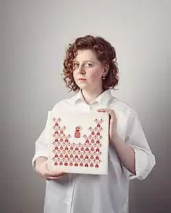 Rufina Bazlova ist stehend abgebildet. Sie hält ein quadratisches Kunstwerk, auf dem rote Piktogramme auf weißem Untergrund zu sehen sind. Bazlovas rotes lockiges Haar ist halblang und sie trägt eine weite weiße Bluse. Sie schaut direkt in die Kamera.