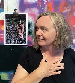 Ruth Bamberg mit schulterlangen, grauen Haaren in schwarzem Shirt, die nachdenklich zur Seite schaut. Im Hintergrund ein Foto eines nackten Mannes, der auf Steinen sitzt.