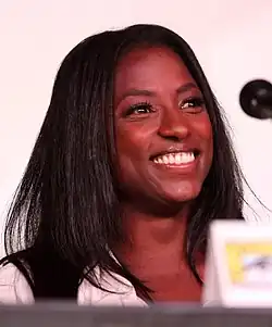 Rutina Wesley lächelt während einer Panel-Diskussion auf der Comic-Con 2012 in San Diego. Sie hat langes, glattes, schwarzes Haar und trägt ein weißes Hemd. Sie sitzt an einem Tisch, auf dem ein Mikrofon sichtbar ist. Vor ihr befindet sich ein Namensschild mit dem Comic-Con-Logo, das die Veranstaltung kennzeichnet.