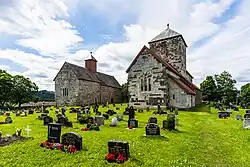 Foto zweier nebeneinandergelegenen, ähnlich aussehenden steinernen Kirchen, wobei die rechte Kirche etwas größer ist. Im Vordergrund befinden sich Grabsteine.