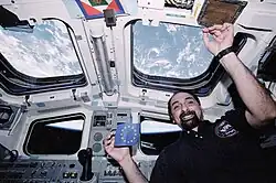 Astronaut Guidoni posiert mit einer europäischen Flagge vor einem Fenster des Space Shuttle, im Hintergrund ist die Erde zu erkennen.