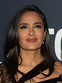 Salma Hayek, 2024 auf dem Toronto International Film Festival mit langen, dunklen Haaren, die locker über die Schultern fallen. Sie trägt ein elegantes schwarzes Kleid mit einem asymmetrischen Ausschnitt, der diagonal über die Schultern verläuft. Der Hintergrund ist dunkel gehalten, wobei rechts neben der Person ein Fragment eines weißen Buchstabens zu sehen ist. Dieses Fragment könnte zu einem Schriftzug oder Logo gehören, ist jedoch nicht vollständig erkennbar. Der Fokus des Bildes liegt auf der Person und ihrem Outfit, wobei der dunkle Hintergrund eine gewisse Tiefe und Kontrast bietet.
