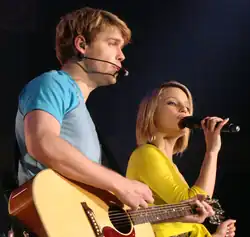 Dianna Agron im Duett mit Quinn Fabray auf dem Glee Live-Konzert 2011. Er links trägt ein blaues Hemd und spielt eine Akustikgitarre. Dianna Agron rechts trägt ein gelbes Oberteil und hält ein Mikrofon, was darauf hinweist, dass sie singt. Das Bild erfasst einen Moment einer Live-Performance und hebt die Zusammenarbeit zwischen dem Gitarristen und der Sängerin hervor. Die Bühnenbeleuchtung und die Musikinstrumente betonen die Atmosphäre einer musikalischen Darbietung.