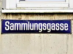 Blaues Straßenschild mit weißer Schrift Sammlungsgasse an einer Wand unterhalb eines Fensterbrettes angebracht