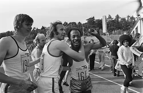 In einer Gruppe von Leichtathleten in Trikots umarmt ein europäischer Athlet mit Locken einen afroamerikanischen Athleten mit Kraushaar. SChwarz-weiß-Fotografie aus Papendal 1975.
