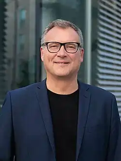 Sascha Adamek, 2023. Brille, kurze Haare blaues Jacket und schwarzes T-Shirt, freundliches Lächeln