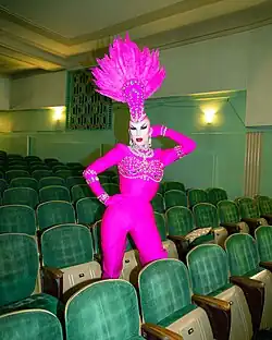 Velour steht inmitten einer Reihe hochgeklappter beiger Sitzbänke mit braunen Armlehnen und dunkelgrün-weiß kariertem Bezug. Sie trägt roten Lippenstift, rosa Lidschatten, Wimperntusche, falsche Wimpern, aufgemalte Augenbrauen, ein lila Kostüm mit Edelsteinen an der Brust und den Ärmeln sowie eine Halskette und Ohrringe aus Diamanten. Auf ihrem kahl rasierten Kopf befindet sich eine längliche Kopfbedeckung mit Edelsteinen und nach oben abstehenden Federn. An der dunkelgrünen Wand hinter ihr befinden sich zwei runde, gelb leuchtende Lampen und ein türkis-schwarz gestreifter Teppich mit braunem Bogenmuster.