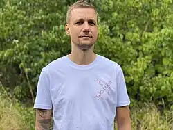 Tobias Schlegl steht im Grünen, bekleidet mit einem weißen T-Shirt, auf dem das Logo seines Romans „Schockraum“ abgedruckt ist.