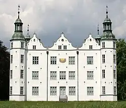 Frontale Farbfotografie eines dreigeschossigen Schlosses mit einer Sonnenuhr im Obergeschoss und vier Ecktürmen, auf denen Turmhauben sind. Zwischen den vorderen Türmen befinden sich drei Staffelgiebel. Im Vordergrund steht eine Wiese und an beiden Seiten sind Bäume zu sehen.