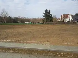 im Vordergrund oberste Treppenstufen der Freitreppe. Im Blickfeld ein ebener Schotterplatz, dahinter ein Grünstreifen, Bäume und Häuser