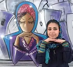 Shamsia Hassani nimmt dieselbe Pose ein, wie ihre Graffiti-Figur im Hintergrund: verschränkte Hände unter dem Kinn. Hassani trägt ein blaues Kopftuch und ihr dunkles Haar ist leicht zu sehen. Das Graffiti zeigt eine Frau mit rotem Kopftuch und lilafarbener Kleidung.