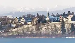 Tele-Aufnahme der Gemeinde Fehraltorf. Auf einem Hügel stehen einige Wohngebäude sowie die Kirche. Direkt unterhalb ist knapp der Pfäffikersee erkennbar.