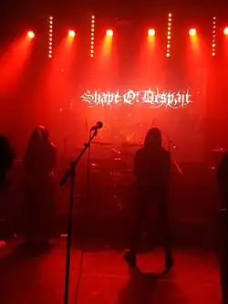 Sängerin und Sänger stehen mit tief gezogenen Kapuzen auf einer rot beleuchteten Bühne vor dem Schlagzeug.