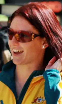 Shayne Reese im gelb-grünen Trainingsanzug lächelt, trägt eine Sonnenbrille und streift mit der linken Hand ihr Haar zurück.