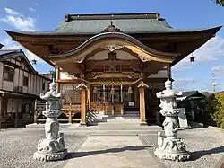 Frontalansicht eines kleinen Shintō-Schreins. Auf dem Weg zum Schrein befindet sich links und rechts eine weiße Steinsäule, die jeweils mit sie umwindenden Schlangen verziert ist.