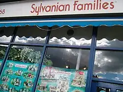 Über einem Schaufenster steht auf weißem Hintergrund die rote Aufschrift Sylvanian Families. Im Schaufenster sind einige Leuchten, Pflanzen sowie Poster mit verschiedenen Tier-Spielfiguren und Puppenhäusern zu sehen. Im Schaufenster spiegelt sich der mit grauen Wolken bedeckte Himmel.