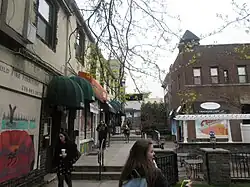 Gehweg zwischen beidseitiger, zweigeschossiger Bebauung, ansteigendes Gelände mit Treppen, ein halbes Dutzend Personen im Bild gehen ihrem Alltag nach. Links zweigeschossige geschlossene Reihenbebauung mit Geschäften und Eingangstüren, im Obergeschoss Wohnungen.