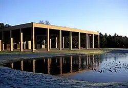 Seitliche Farbfotografie eines hellbraunen, schlichten Gebäudes mit rechteckigen Säulenvorhöfen an allen drei Seiten. Die hintere Mauer ist schwer zu erkennen. Im Hintergrund stehen Bäume und im Vordergrund ein Teich mit frostigem Ufer.