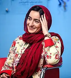 35-jährige Frau auf Stuhl sitzend, freundlich lächelnd, mit Kopftuch, den Kopf leicht mit dem li. Arm abstützend