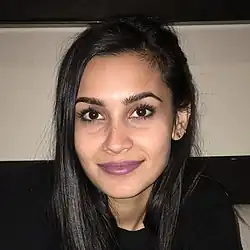 Sophia Ali mit langen, dunklen Haaren und einem freundlichen Lächeln. Sie trägt einen schwarzen Pullover. Auf dem linken Ohr ist ein kleines Ohrstecker-Piercing erkennbar. Der Hintergrund des Bildes scheint sich in einem Innenraum zu befinden. Eine beige Wand und ein dunkler, horizontaler Balken oder ein Regal bilden die Kulisse. Das Licht ist gedämpft, was darauf hindeutet, dass die Aufnahme bei schwachen Lichtverhältnissen gemacht wurde.