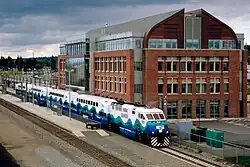 Sicht von schräg oben auf Bahnhof mit zwei Gleisen an einem Mittelbahnsteig. Ein Doppelstock-Wendezug mit Lokomotive unten in Bildmitte und vier nach links hinten sichtbaren Doppelstockwagen steht an der abgewandten Bahnsteigkante. Der ganze Zug ist einheitlich im Sound-Transit-Farbschema lackiert: Weiß am Dach und Oberdeck, nach unten hin wie Meereswellen in drei Farbabstufungen Türkis, Dunkelgrün, Dunkelblau. Die rechte Bildhälfte dominiert ein viergeschossiges, modernes Bahnhofs- oder Bürogebäude mit rötlicher Klinkerfassade und Glasflächen sowie einem schmetterlingsartig geschwungenen dunklen Dach. Dräuend bewölkter Himmel, Wald und eine Hauptstraße im Bildhintergrund.