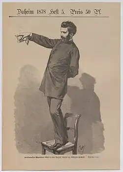 Ein junger bärtiger Mann steht auf einem Stuhl, eine Hand hinter dem Rücken, die anderen mit ausgestreckten Zeigefinger nach links weisend – in dieser Hand hält er auch ein Taschentuch o.&nbsp;ä.