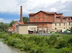 altes Firmengebäude aus Backstein. Im Vordergrund das Ufer eines Gewässers