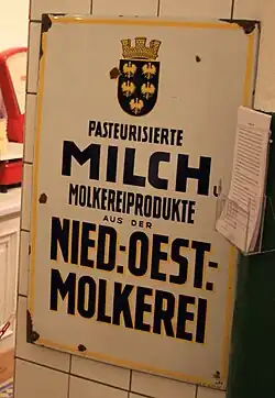 Emailleschild, weißer Hintergrund, gelb umrahmt mit schwarzer Beschriftung „Pasteurisierte Milch und Molkereiprodukte aus der Niederösterreichischen Molkerei“