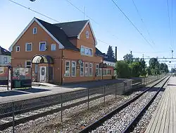 Foto eines Bahnhofsgebäudes mit Schienen im Vordergrund. Das Gebäude ist mit orange gestrichenem Holz verkleidet.