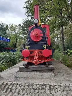 Es ist eine rot, schwarz, gelb bemalte Schmalspurdampflok auf einem Sockel vor dem Bahnhof der Kindereisenbahn zu sehen.