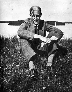 Mit angewinkelten Beinen im Gras, welches dahinter an eine Steinfläche angrenzt, sitzender Mann mittleren Alters im grauen Anzug, hellem Hemd, Krawatte, grauen Socken und schwarzen Schuhen mit Blick in die Kamera in schwarzweiß.