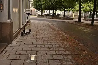 Foto einer Straße und eines Gehwegs mit drei goldenen Markierungen im Boden vor einem Hauseingang. Auf dem Gehweg befindet sich zudem ein E-Roller