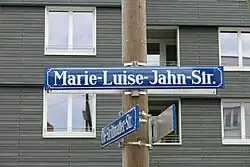 Blaues Straßenschild mit weißer Schrift „Marie-Luise-Jahn-Str.“. Das Schild ist an einem runden Holzpfahl angebracht, dahinter sieht man ein graues Gebäude.