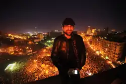 Sultan Sooud Al-Qassemi oberhalb des Tahrir-Platzes in Kairo, Ägypten, 2012. Ein arabischer Mann mit Ballonmütze vor dem dunklen Hintergrund eines erleuchteten Platzes bei Nacht.