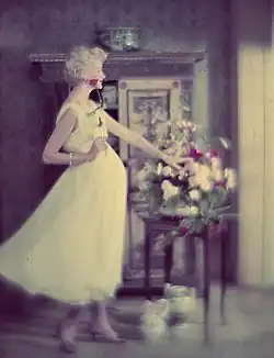 Sunny Harnett in einem weißen Kleid, die in der Hand eine einzelne Blume hält. Neben ihr steht ein Tisch, auf dem ein Blumenstrauß kunstvoll arrangiert wurde. Im Hintergrund befindet sich ein antikes Möbelstück mit dekorativen Elementen, auf dem eine kleine Schale platziert ist. Der Raum ist in gedämpftem Licht gehalten und die Wände sind mit einer gemusterten Tapete versehen, die eine klassische Atmosphäre schafft.