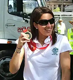 Suzie Fraser trägt ein weißes T-Shirt und eine Sonnenbrille. Die Medaille ist an einem roten Band befestigt. Im Bildhintergrund ist ein Truck zu sehen, daneben stehen zwei Ordner.
