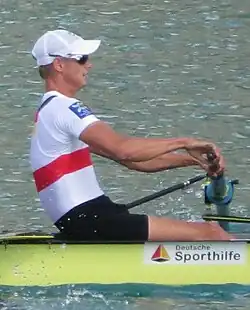 Sven Keßler ist von der Seite im gelben Boot zu sehen. Er trägt eine weiße Mütze, eine Sonnenbrille, das Nationaltrikot und eine schwarze Hose. Sein Ruder befindet sich auf der dem Betrachter abgewandten linken Seite des Bootes.