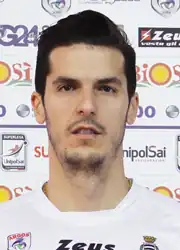 Mann in den zwanziger Jahren im weißt Sporttrikot ab Schulterblatt von vorn vor einer Tafel mit Sponsorenemblemen in die Kamera schauend und in Farbe.