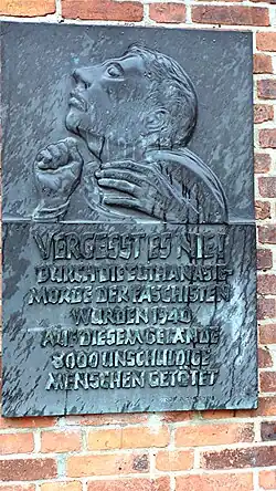 An einer Backsteinmauer ist eine rechteckige Tafel aus dunkelgrauem Metall angebracht. Ihr Relief zeigt eine Person, im seitlichem Porträt mit geschlossenen Augen und dem Gesicht gen Himmel neigend. Die Hände der Person sind zu sehen. Die Rechte Hand ist zur Faust geballt, die Linke schwebt in der Luft. Darunter beginnt die Inschrift.