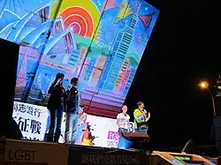 Zu sehen ist Josephine Ho aus der Ferne, sie steht auf einer Bühne die bunt beleuchtet ist. Links im Vordergrund stehen Gebärdendolmetscherinnen.